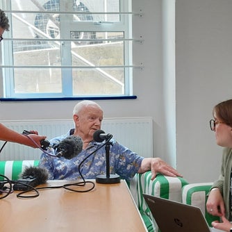 Three people around a table, one is holding a micophone, one is asking questions and the third is answer as the audio trail is recorded
